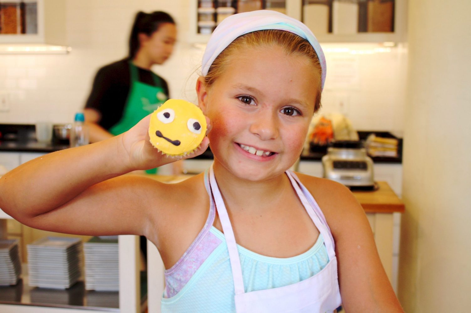 a girl smiling holding up a straight faced emoji cupcake