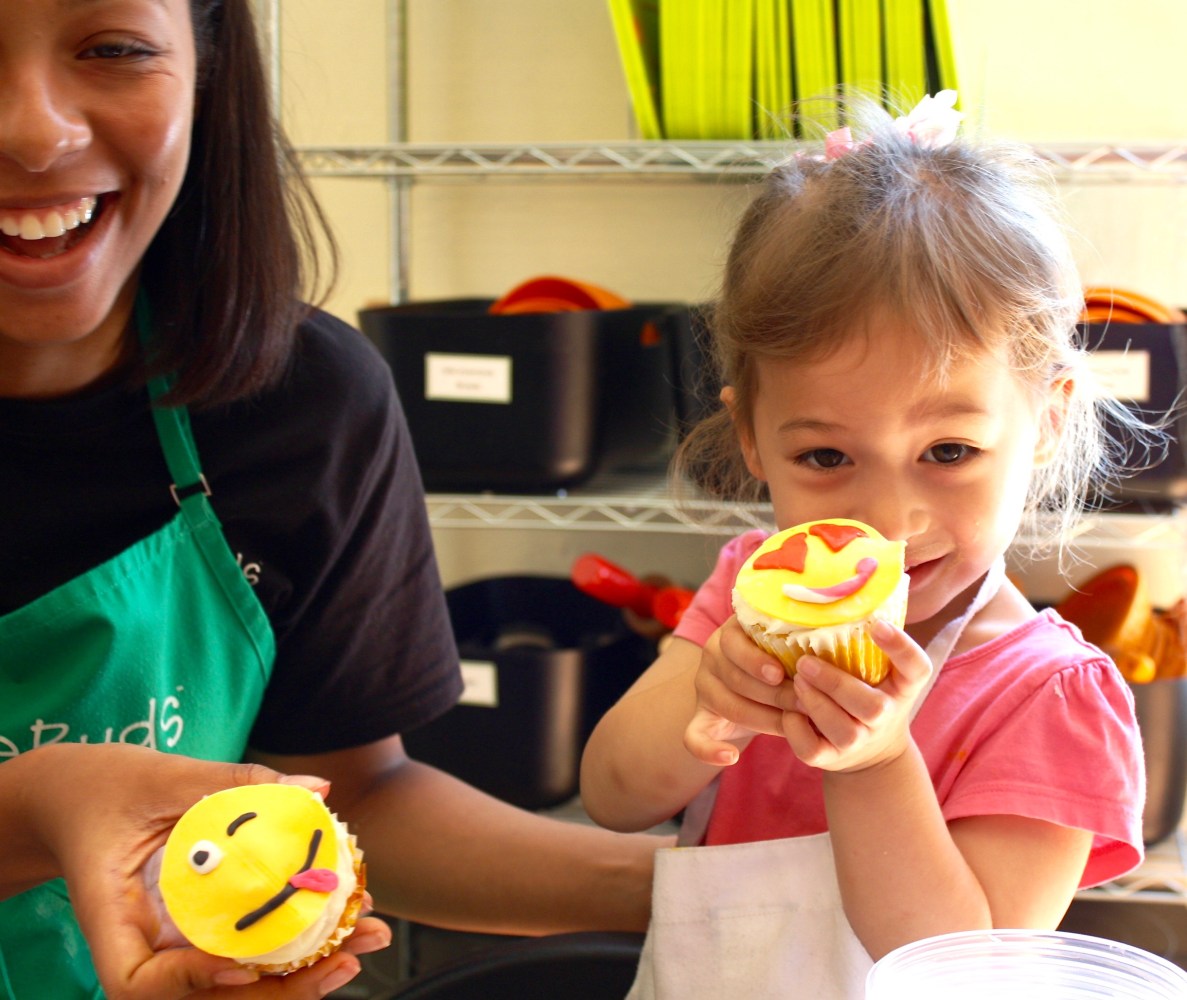 cooking instructor and little girl holding up different emoji face cupcakes