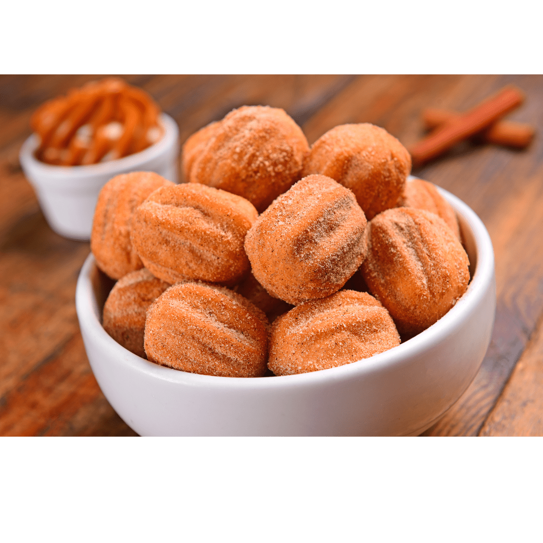 Bowl of small churros dusted with sugar and a bowl of pretzels in the background on a wooden surface.