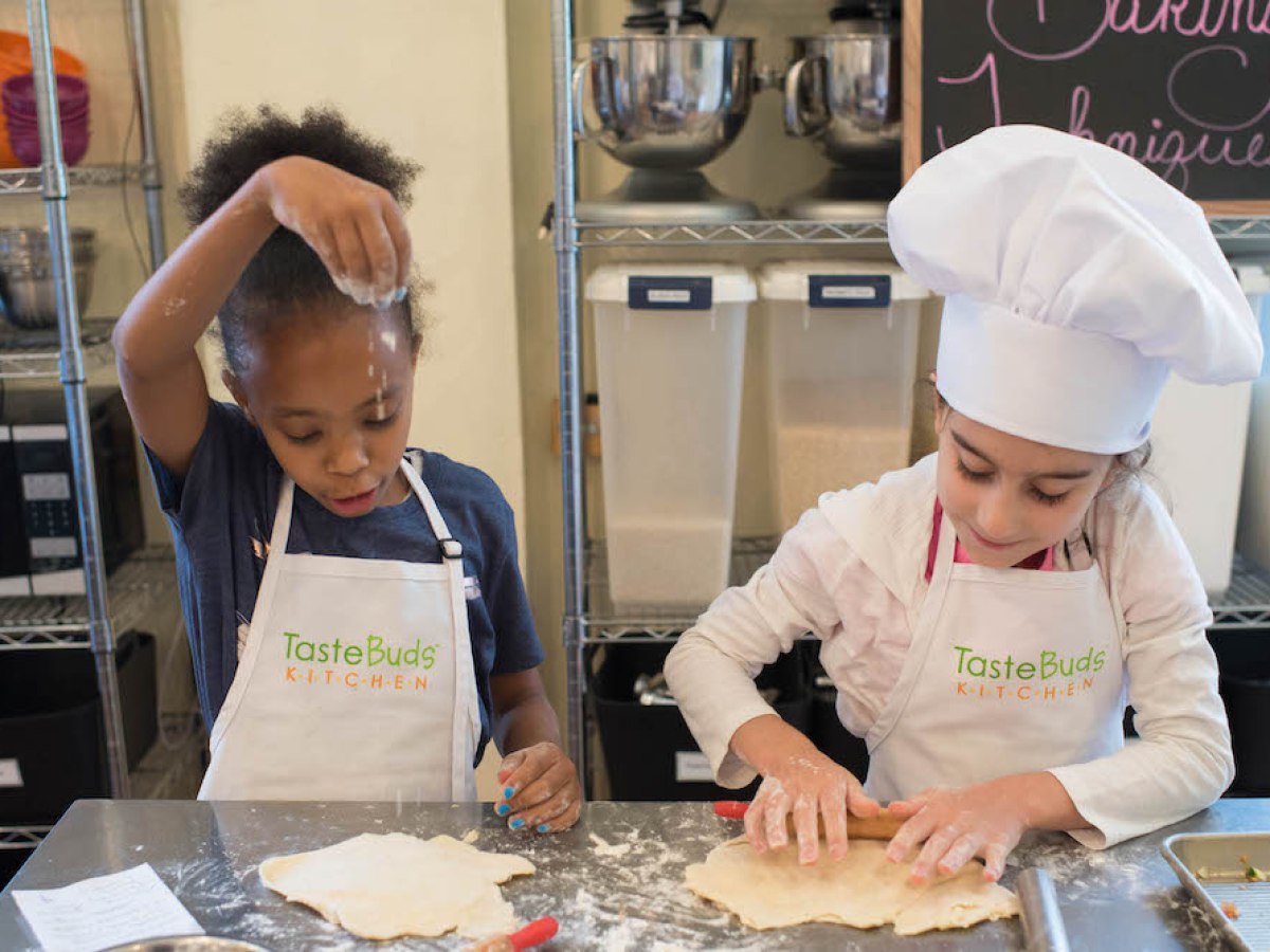 Two children baking with dough; one sprinkles flour, the other rolls dough. Both wear TasteBuds Kitchen aprons.