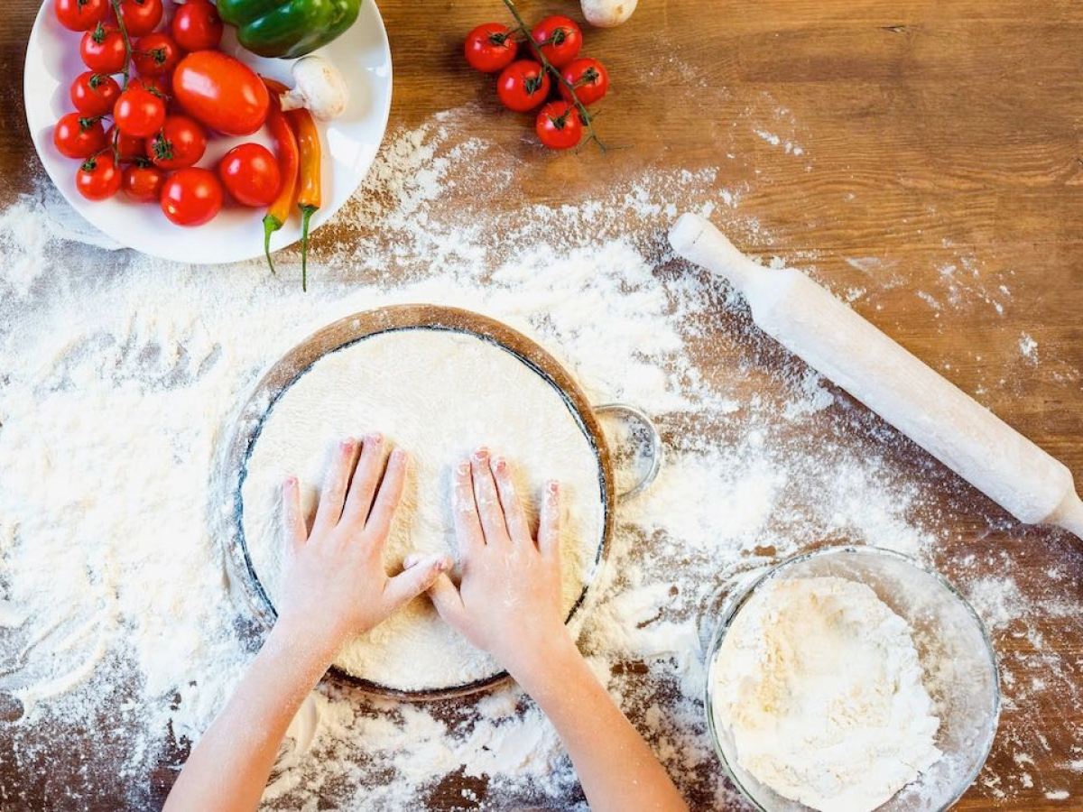 Hands flattening dough with flour, surrounded by a rolling pin and fresh vegetables on a wooden table.