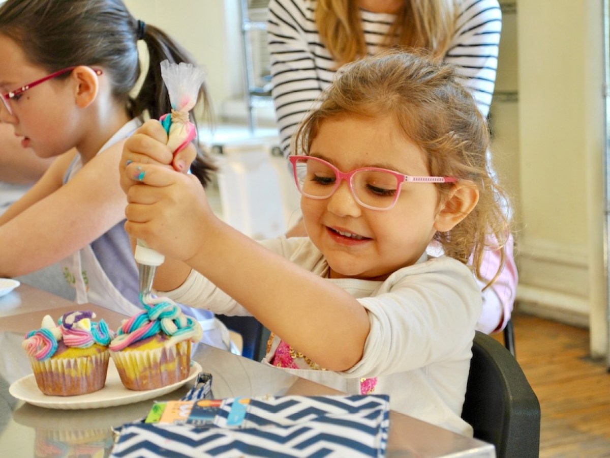 Child smiling while decorating cupcakes with colorful icing at a table.