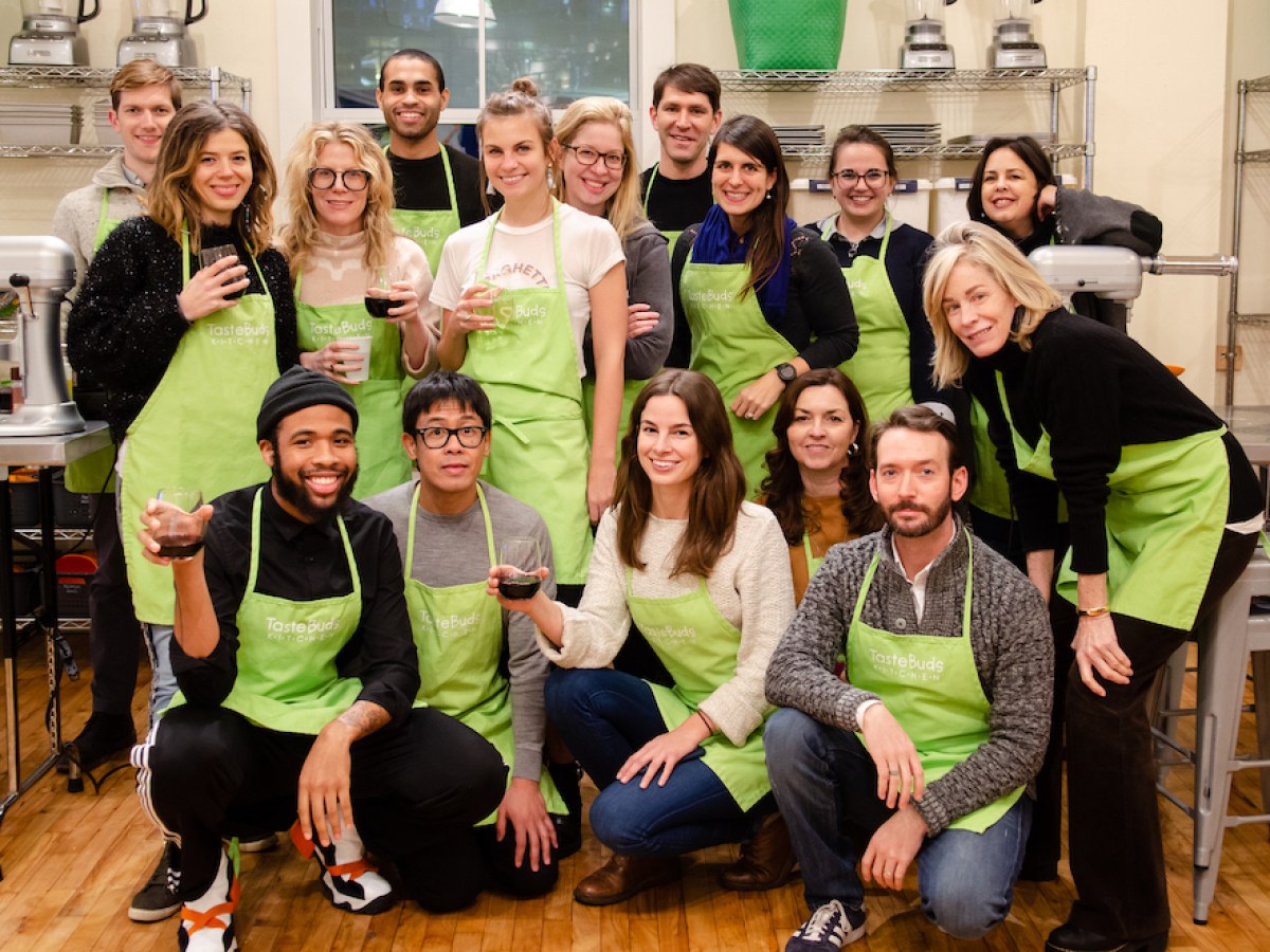 Group of people in green aprons posing with drinks in a kitchen setting.