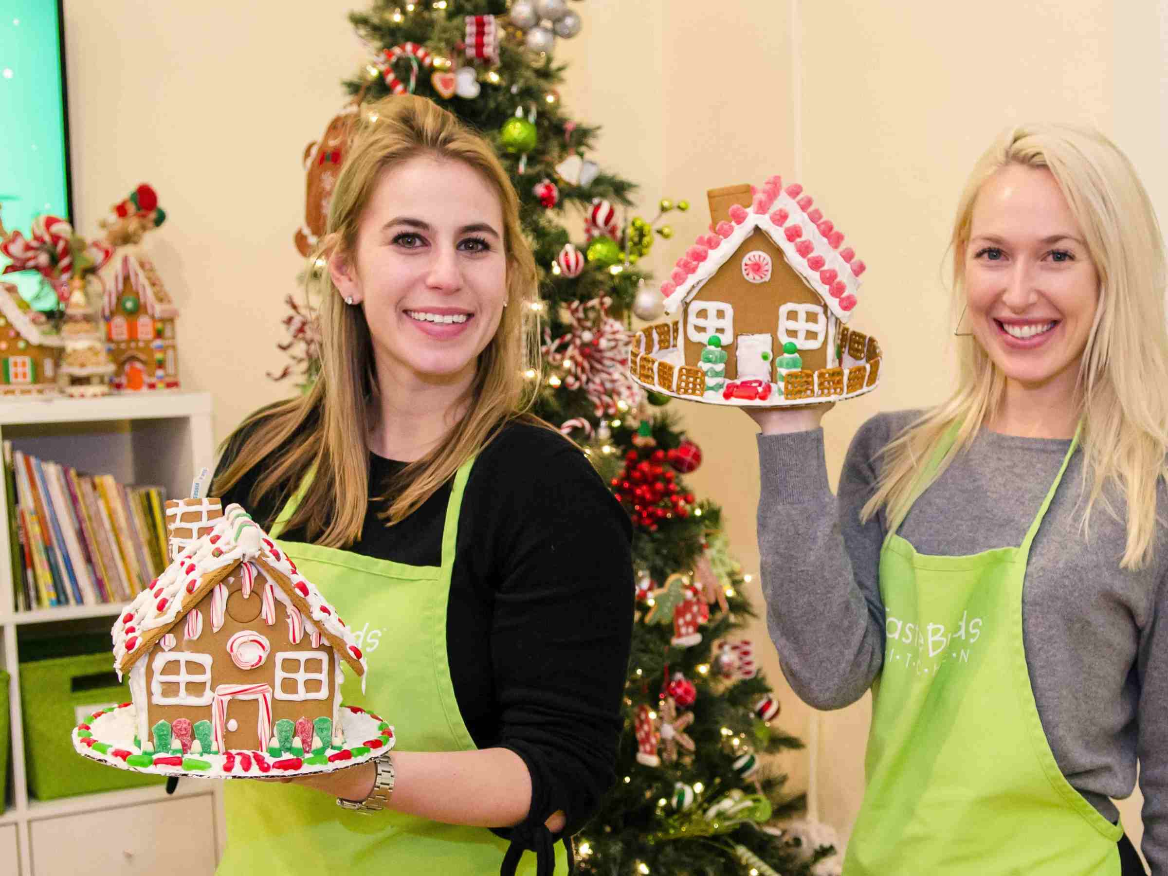 Two women holding gingerbread houses in front of a decorated Christmas tree.