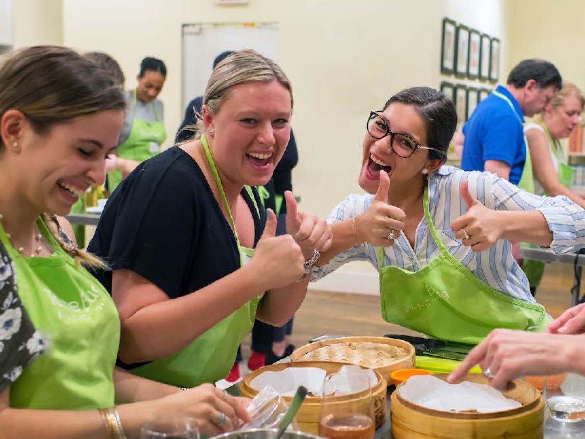 Three women smiling and giving thumbs up while cooking in a class, wearing green aprons.