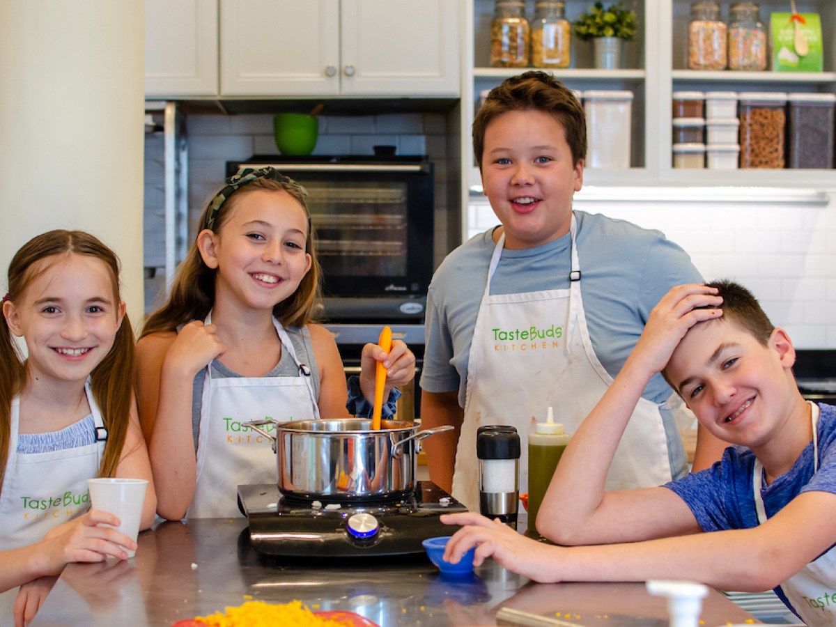 Four smiling kids in a kitchen, wearing aprons, cooking together.